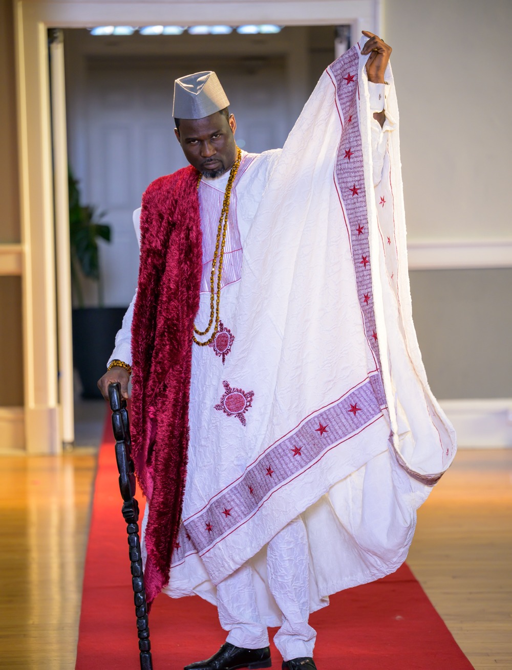 Adeyemi RoyaLabel heritage look, a white ceremonial ensemble with burgundy detailing, traditional cap, beads and carved staff