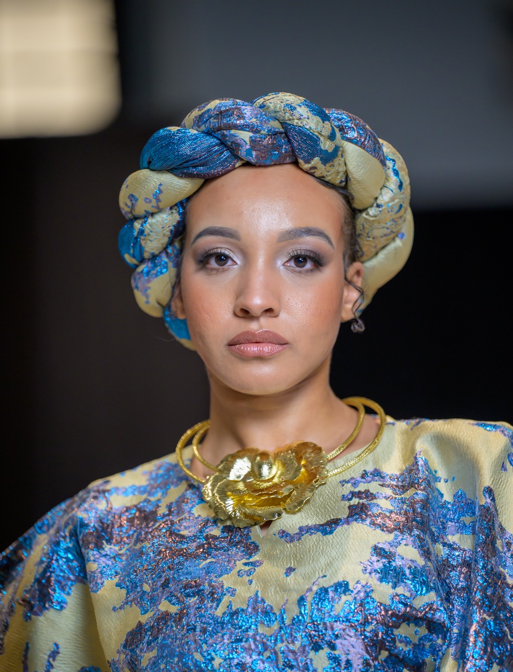 Detail of an Adeyemi Royalabel metallic blue and gold textile with braided headpiece and gold statement necklace